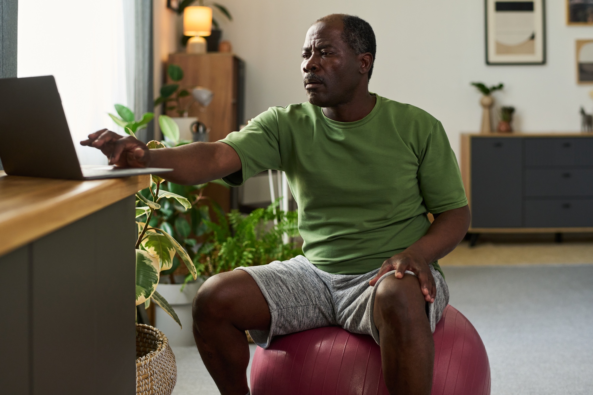 Senior Black Man Sitting on Exercise Ball Using Laptop Indoors